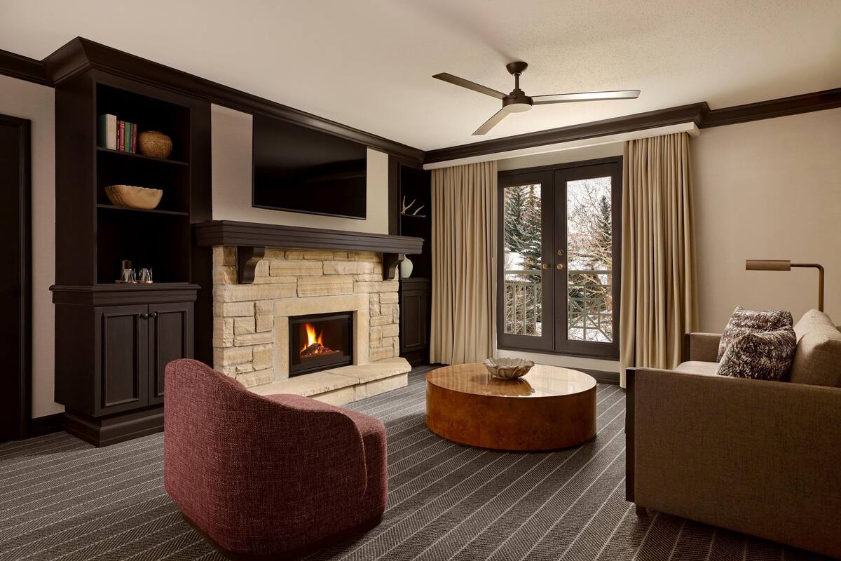 A living room with a fireplace, a ceiling fan, and a large window with curtains.