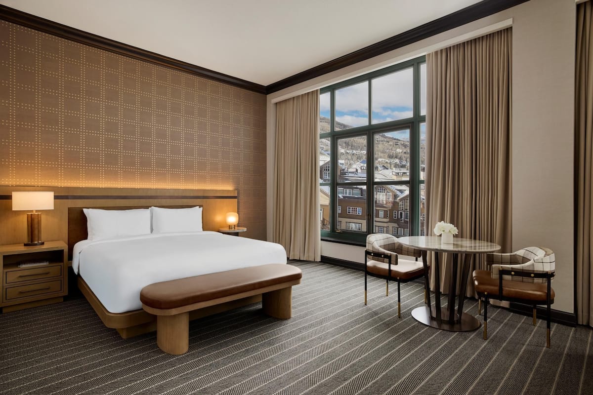 A hotel room with a large bed, a desk with chairs, a window with curtains, and a patterned carpet.