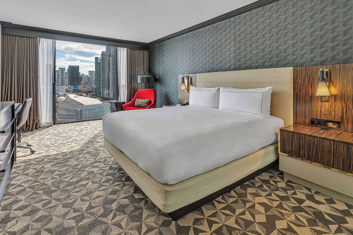 Une chambre d’hôtel avec un grand lit, un fauteuil rouge, un bureau et une vue sur la ville par la fenêtre.