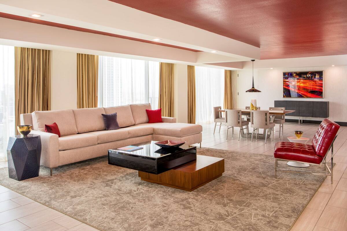 Un salon moderne avec un canapé d’angle beige, une table basse en bois et un fauteuil en cuir rouge. On y trouve également un coin repas avec une table et des chaises blanches, ainsi qu’une grande fenêtre avec des rideaux.