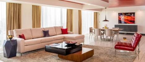 A modern living room with a beige sectional sofa, a wooden coffee table, and a red leather chair. There is a dining area with a white table and chairs, and a large window with curtains.