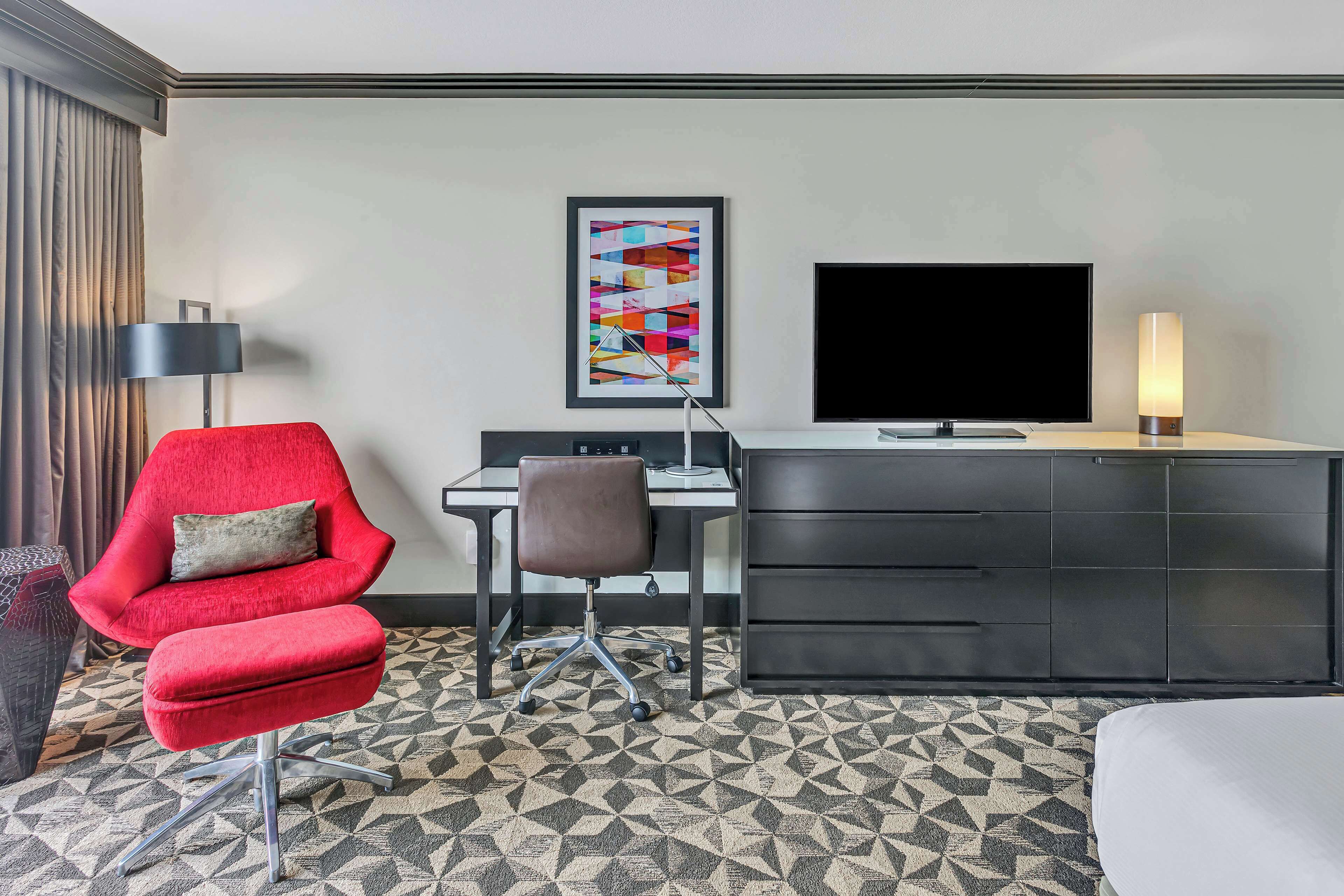 A hotel room with a red armchair, a desk with a computer, a TV, and a cabinet.
