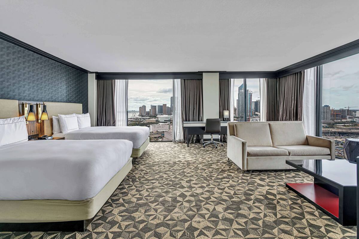 Une chambre d’hôtel avec deux lits, un canapé, un bureau et de grandes fenêtres donnant sur la ville.