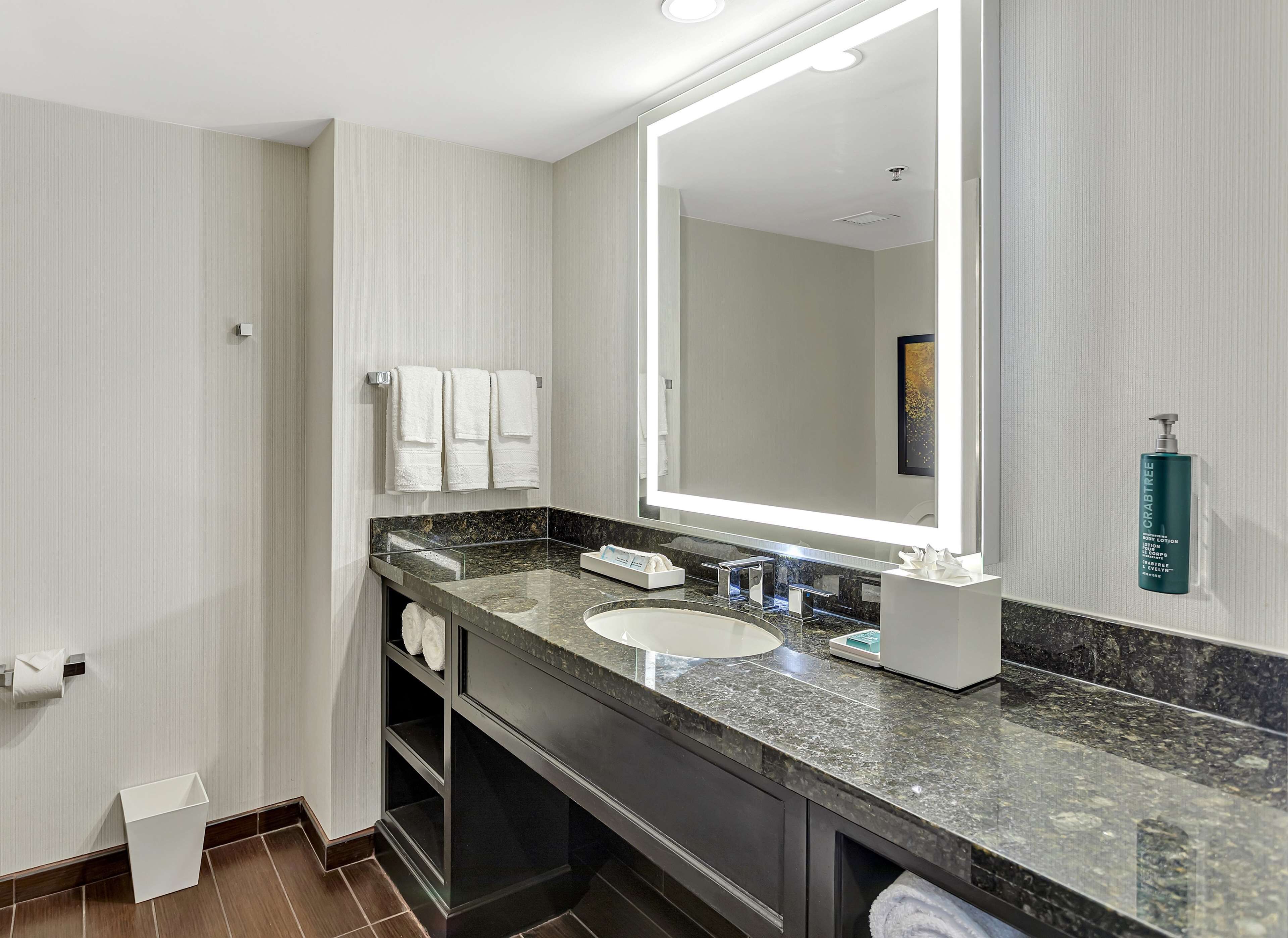 Un bagno moderno con uno specchio grande, un lavandino e un mobile. Asciugamani appesi al muro e una bottiglia di lozione corpo sul ripiano.