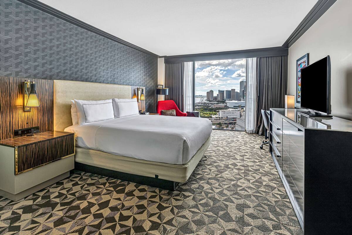 Une chambre d’hôtel avec un grand lit, un fauteuil rouge, un bureau avec une télévision et une vue sur la ville par la fenêtre.