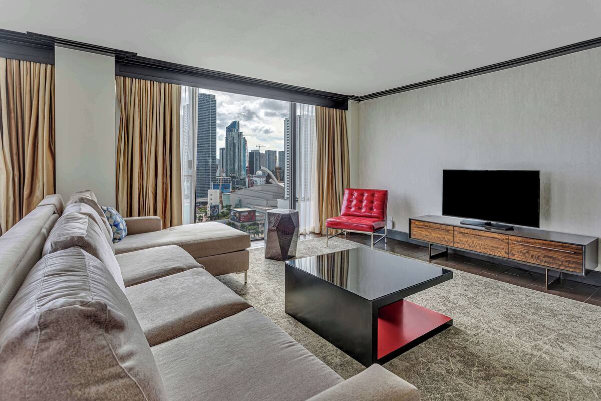 Une chambre d’hôtel moderne avec une grande fenêtre, un canapé, un fauteuil rouge, une télévision et un meuble en bois.