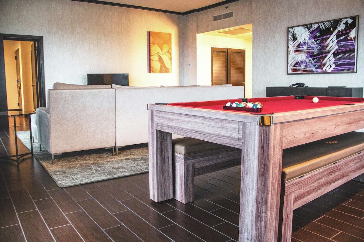 Une table de billard avec des billes, un canapé et un téléviseur dans une pièce dont le sol est carrelé et où l’on aperçoit une porte au fond.