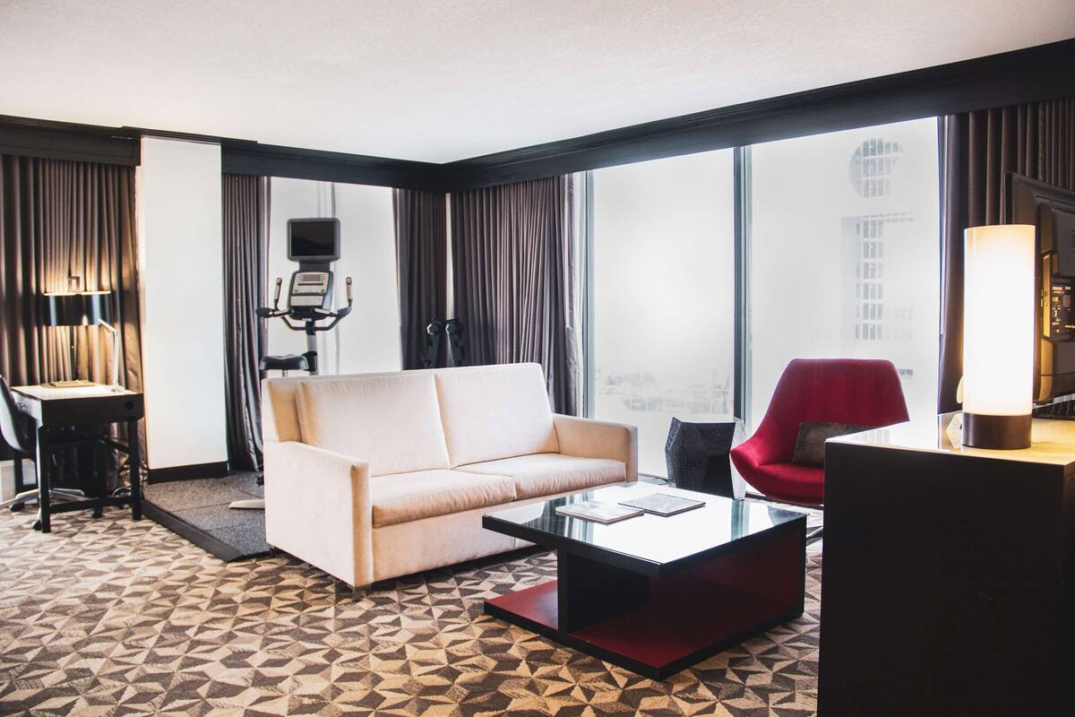 Une chambre d’hôtel avec un canapé, un fauteuil rouge, une table basse, un bureau avec un ordinateur et une grande fenêtre avec des rideaux.