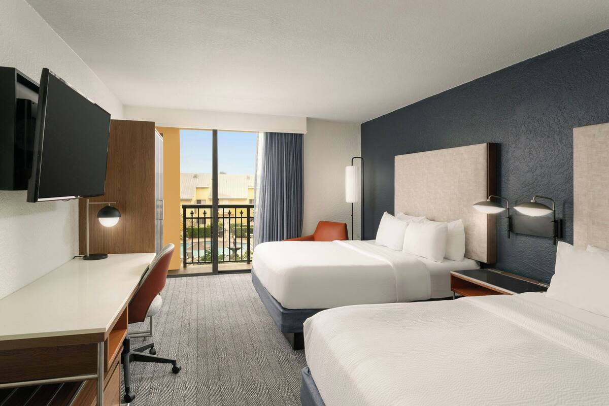 Quarto de hotel com duas camas, uma mesa, uma cadeira, uma TV e uma varanda com vista.