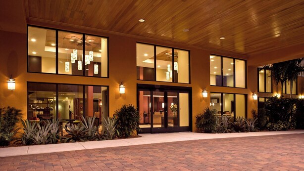 Exterior de un hotel moderno con grandes ventanales, acceso empedrado de ladrillo y asientos al aire libre.