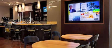 Un restaurante moderno con una zona de bar, mesas de madera y sillas negras. Una pantalla muestra los platos del menú y anuncios.