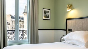 Habitación de hotel con una cama, un cuadro en la pared, una ventana con cortinas y vistas a edificios.