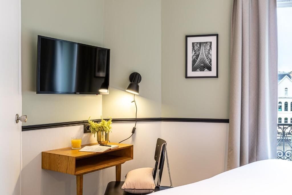 A hotel room with a bed, a desk with a lamp, a TV, and a framed picture on the wall.