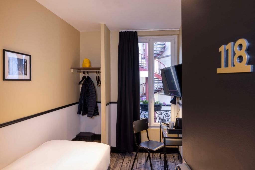 A hotel room with a bed, a chair, a desk with a computer, a TV, and a window with a view of a building and a balcony.