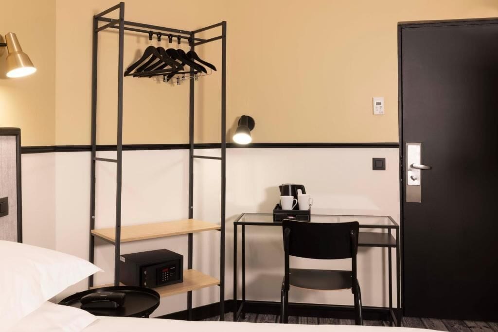 Habitación de hotel compacta con cama, escritorio con silla, estante pequeño, perchero y puerta.