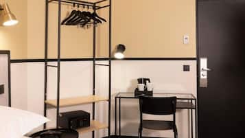 Habitación de hotel compacta con cama, escritorio con silla, estante pequeño, perchero y puerta.