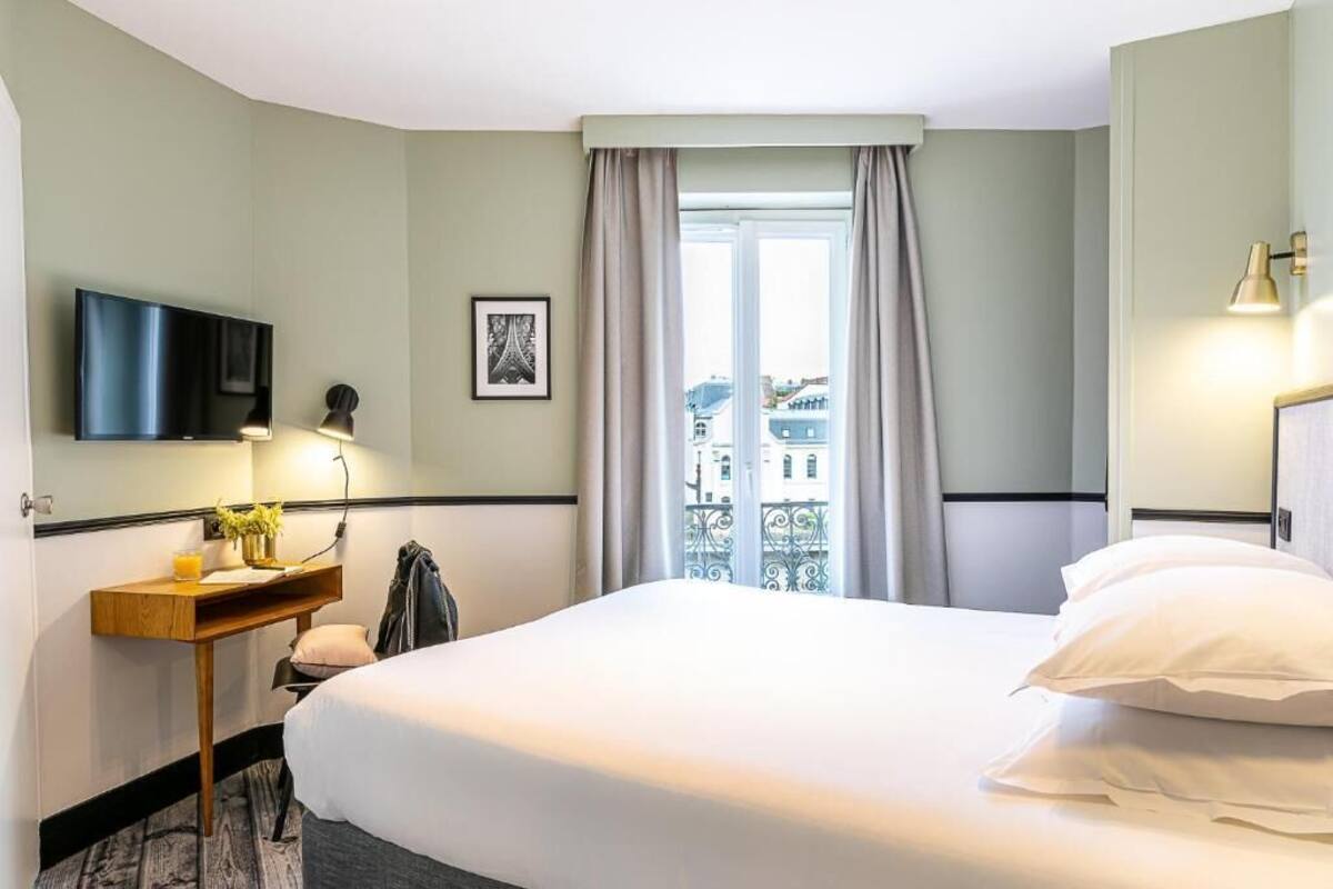 Habitación de hotel con cama, televisor montado en la pared, mesita de noche con lámpara, ventana con cortinas y vista a edificios.