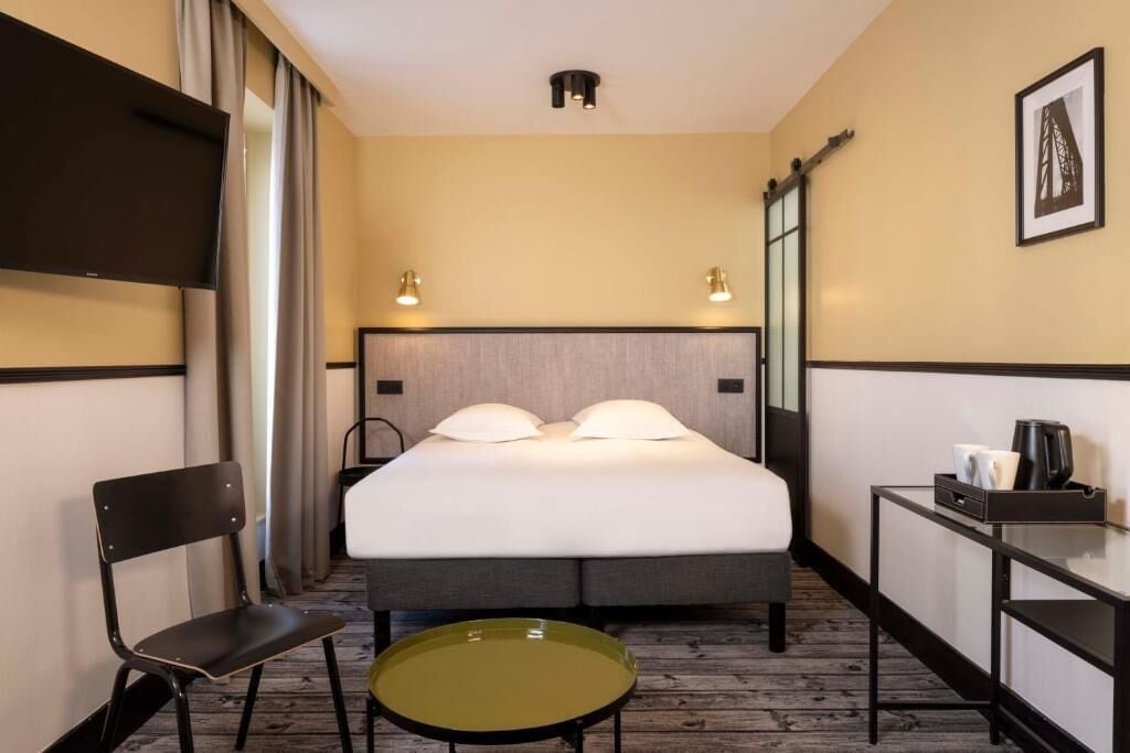 Una habitación de hotel con cama, una silla, una mesita, televisor y cafetera.