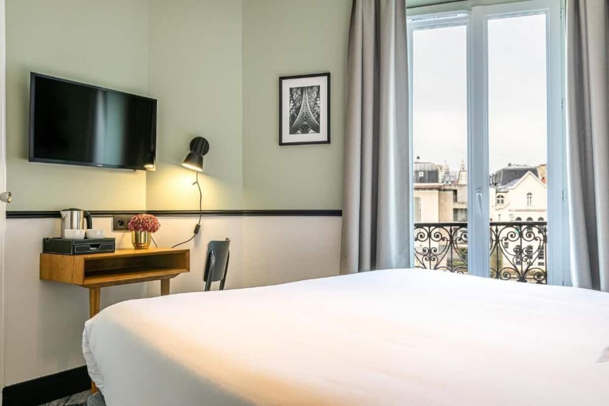 Habitación de hotel con cama, televisor montado en la pared, escritorio con lámpara, cuadro enmarcado y balcón con vistas a edificios.