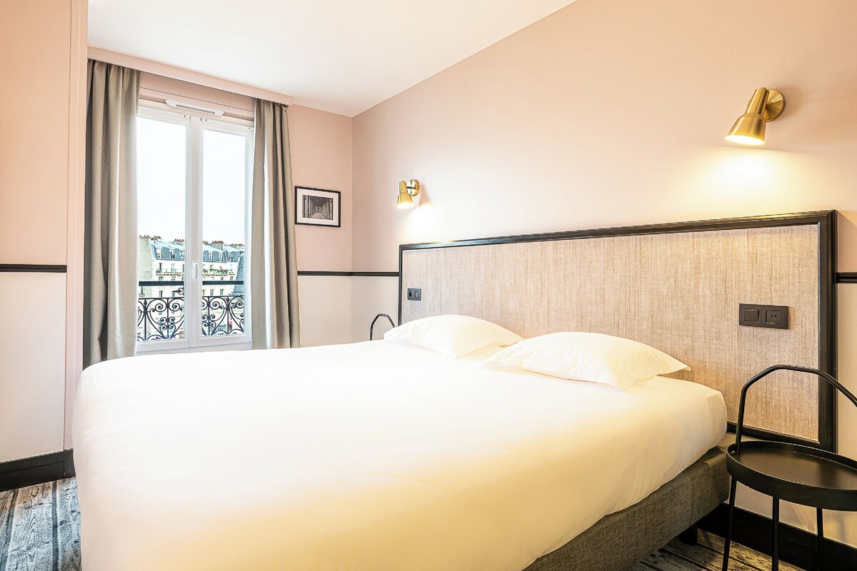 A hotel room with a large bed, a small table, a wall-mounted lamp, and a window with curtains.