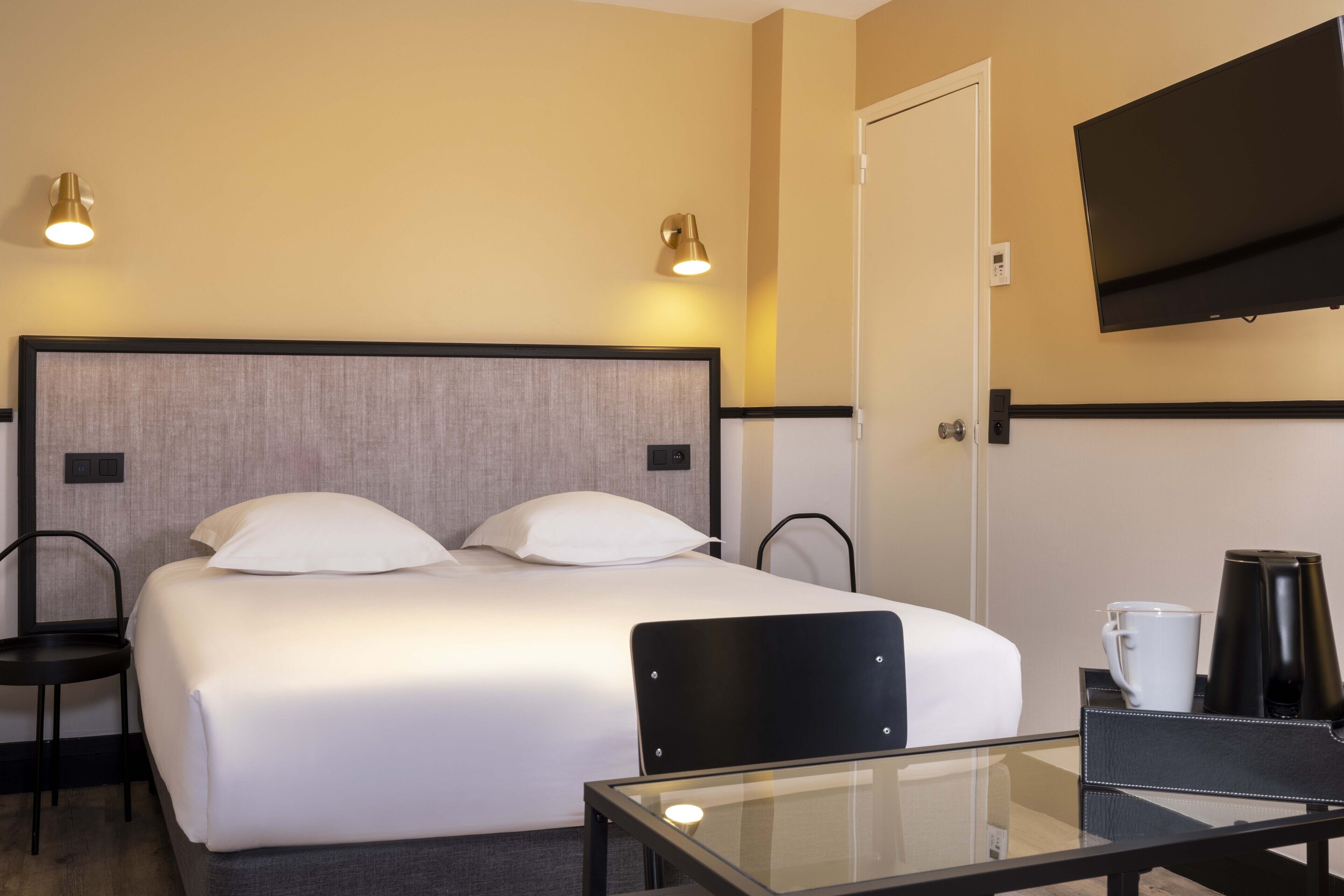 Une chambre d’hôtel comprenant un lit, deux lampes de chevet, un téléviseur fixé au mur, une machine à café et une table en verre.
