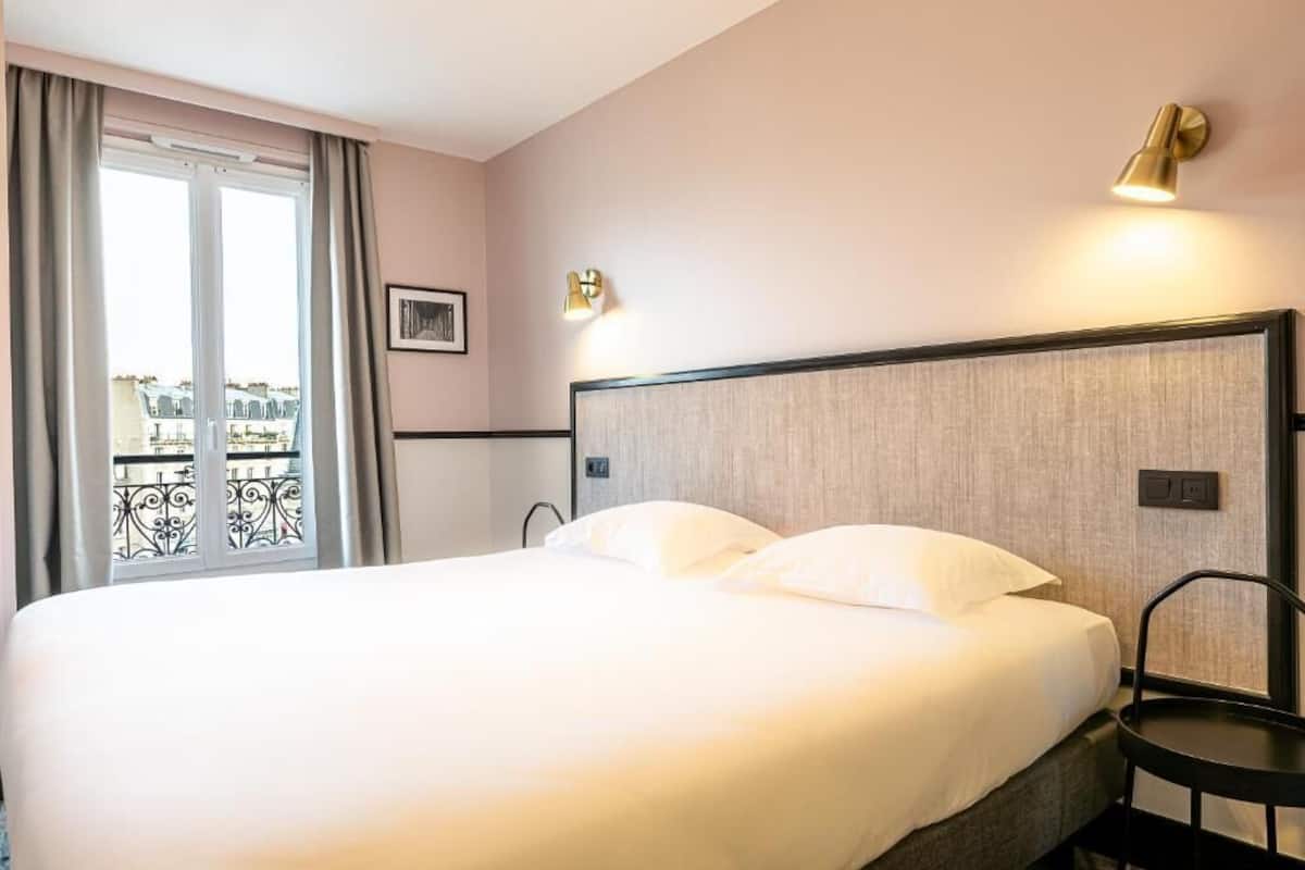Una habitación de hotel con una cama grande, una silla, una ventana con cortinas y una lámpara fijada a la pared.