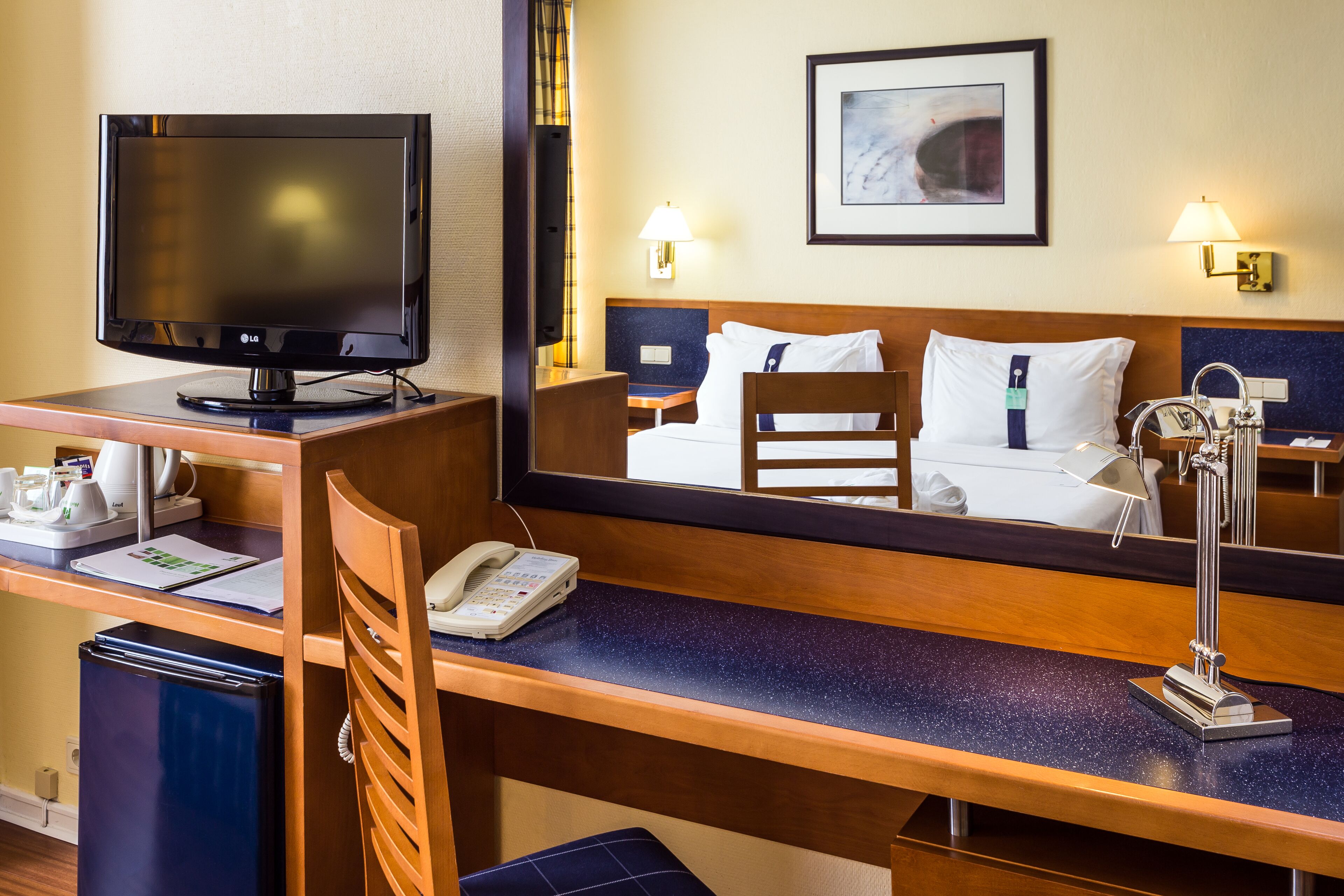 A hotel room with a bed, a desk with a computer monitor, a telephone, and a mirror reflecting the bed.