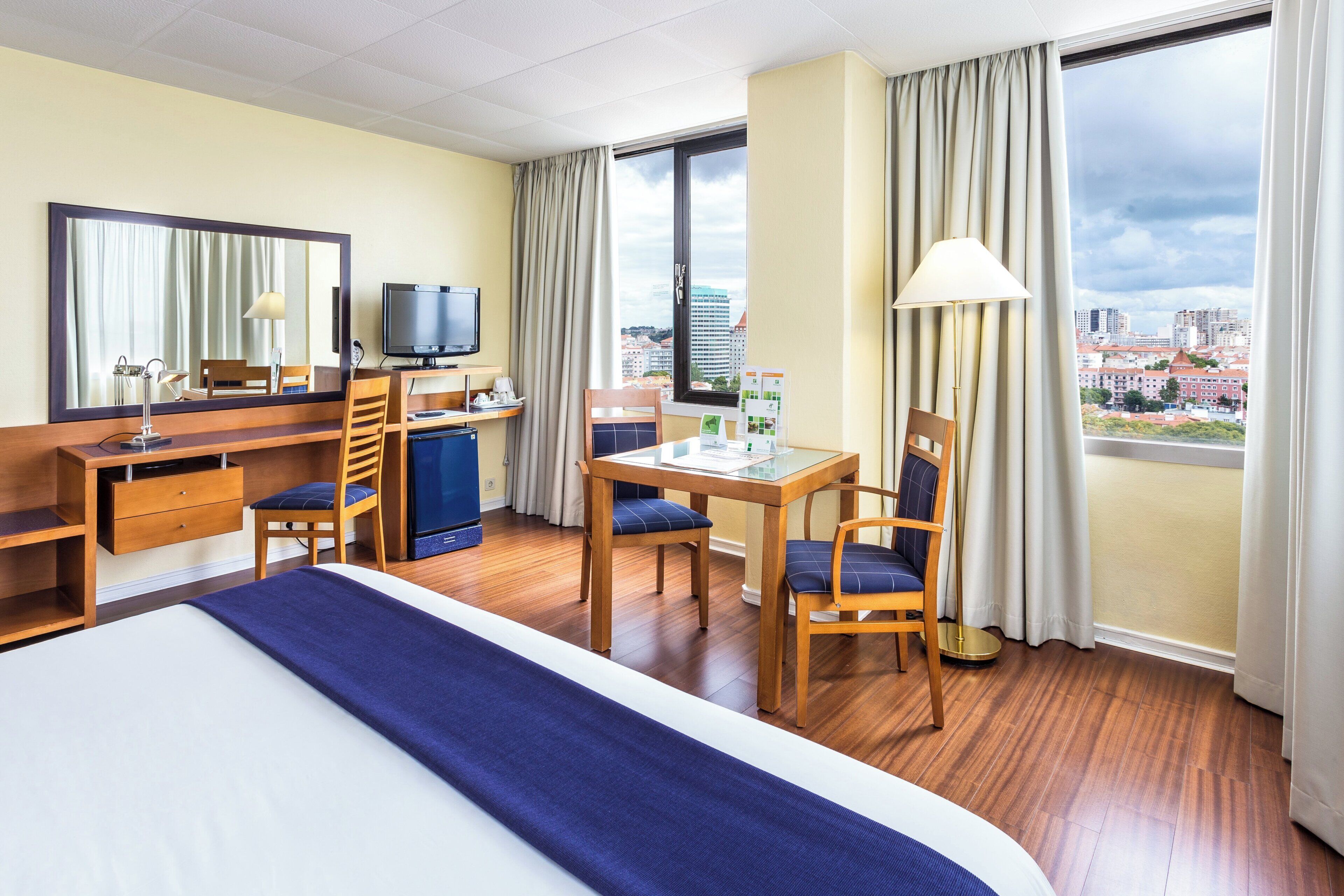 A hotel room with a bed, desk, chairs, a television, and a view of the city.