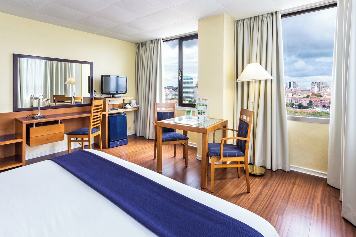 A hotel room with a bed, desk, chairs, a television, and a large window offering a city view.