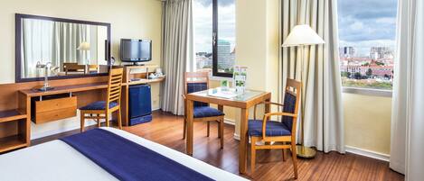 A hotel room with a bed, desk, chairs, a television, and a view of the city.