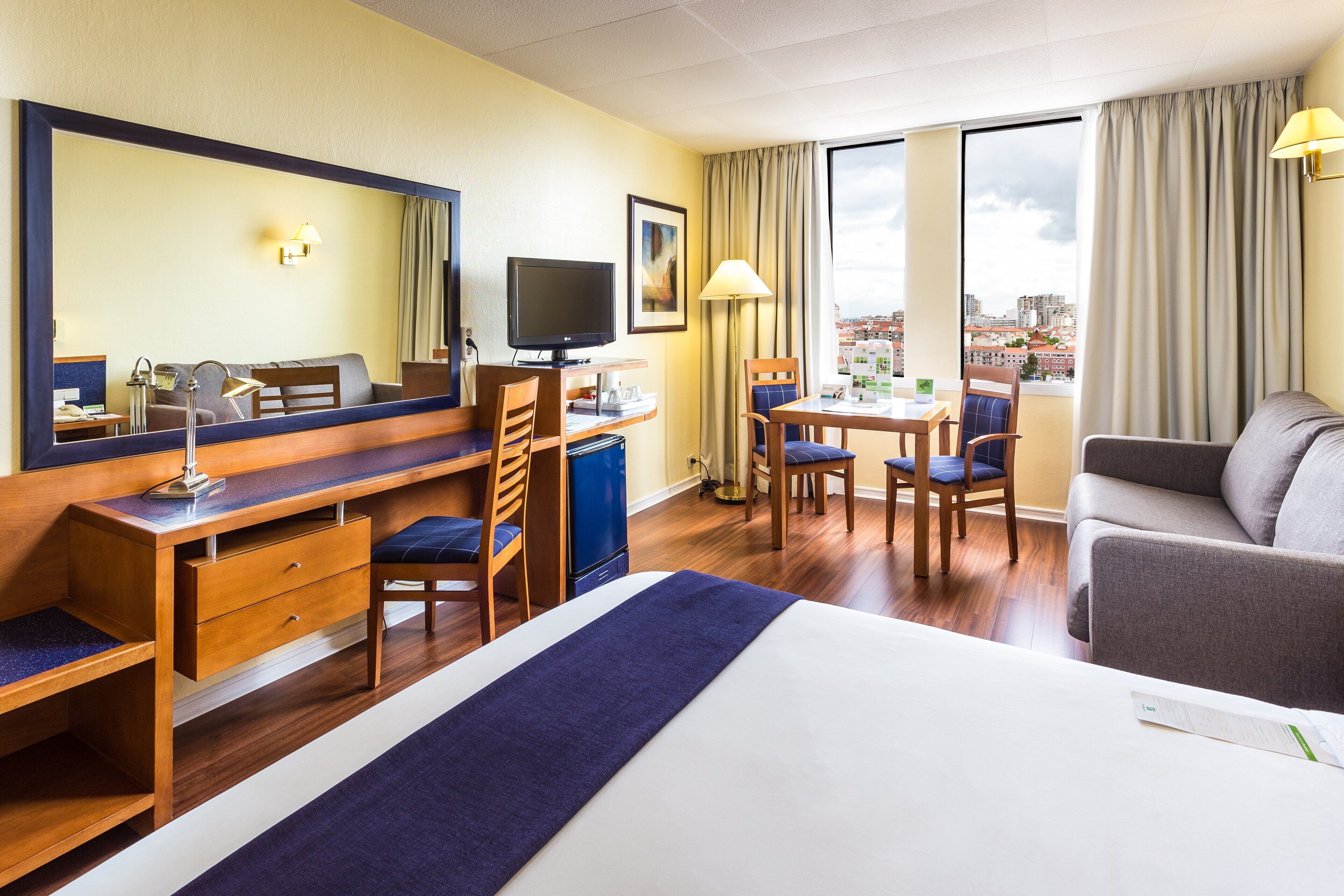 A hotel room with a bed, desk, chairs, a television, and a large window with a city view.