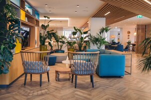 Um hall de hotel moderno com piso de madeira, assentos azuis e diversas plantas.