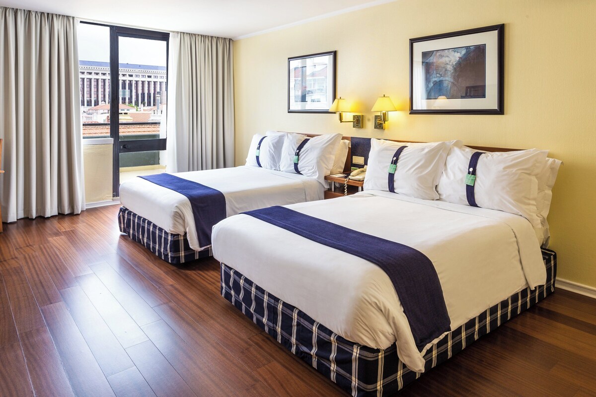 A hotel room with two beds, a view of the city, and a balcony.