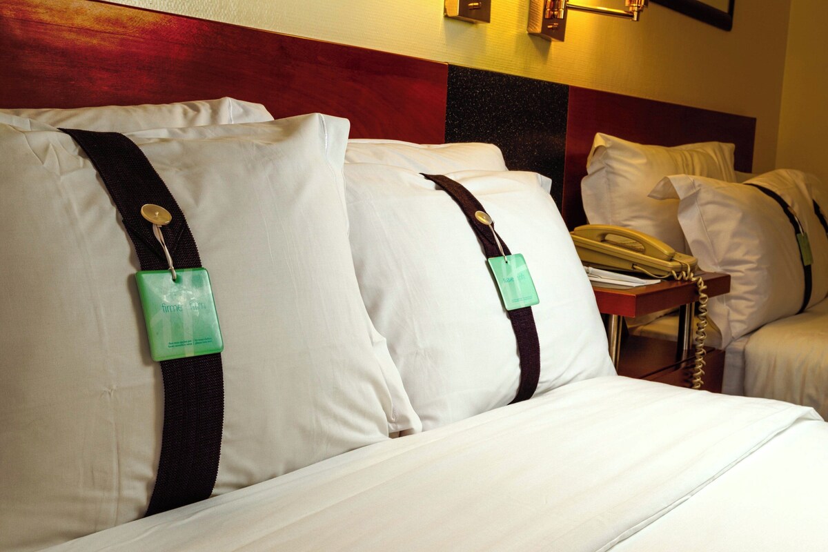 A hotel room with a bed, pillows, a bedside table with a telephone, and a wall-mounted light fixture.