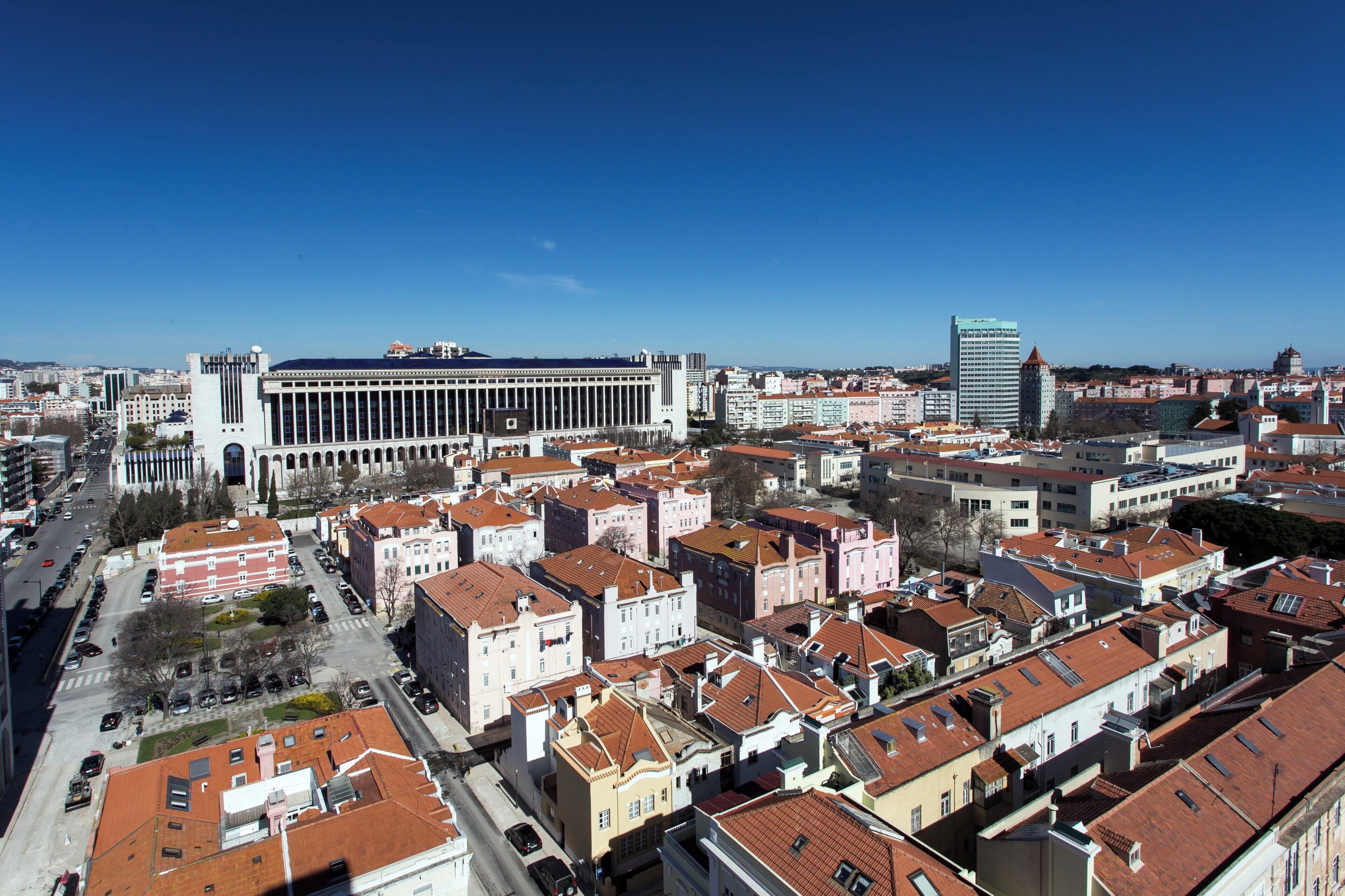 Paisagem urbana com um edifício branco imponente e colunas, cercado por edifícios residenciais com telhados de telha vermelha.