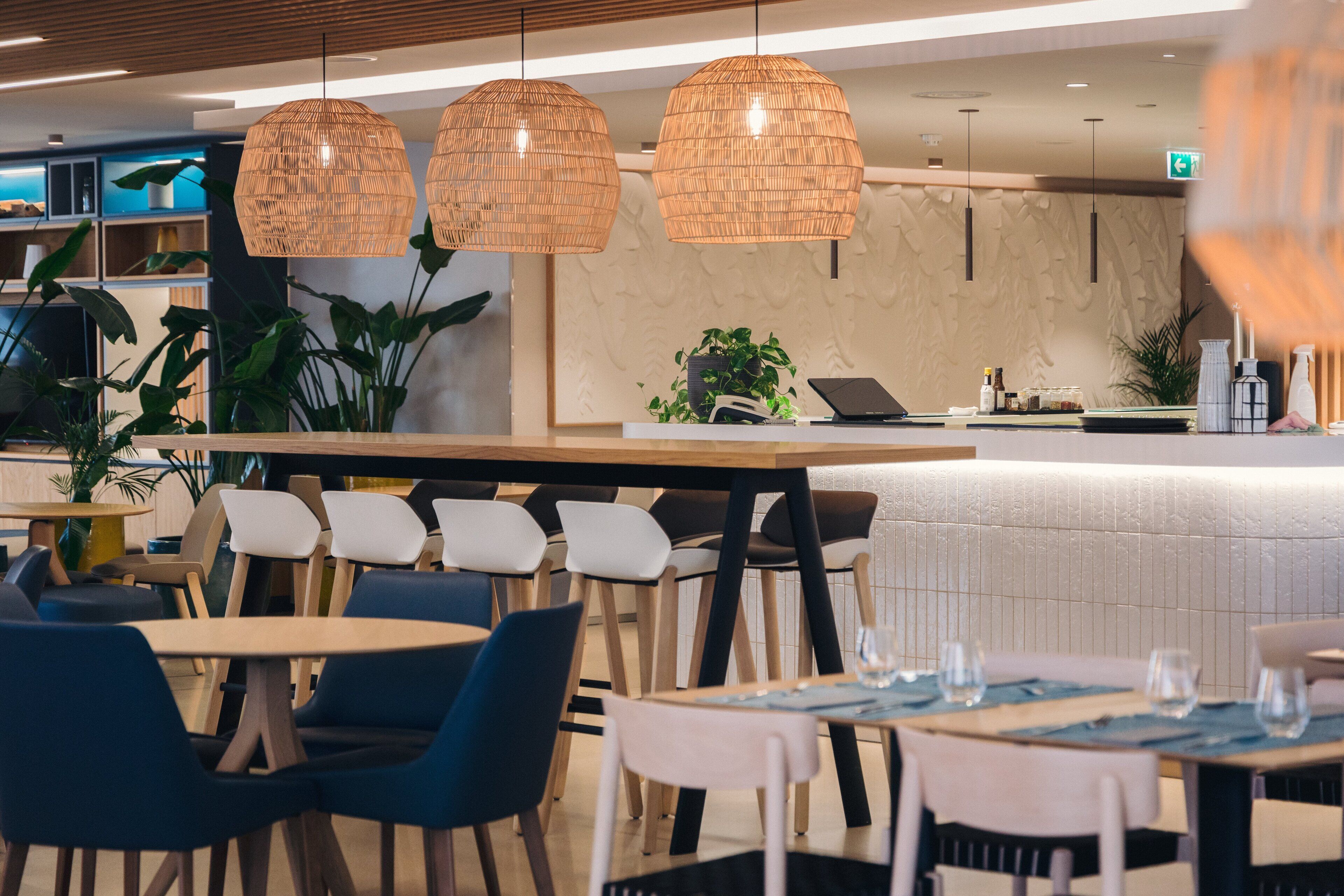 A modern restaurant interior with wooden tables and chairs, hanging wicker lamps, and a counter with a laptop and various items.