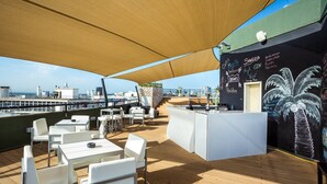 Eine Terrasse im Freien mit weißen Tischen und Stühlen, ein Barbereich mit Kreidetafelmenü und Blick aufs Meer.
