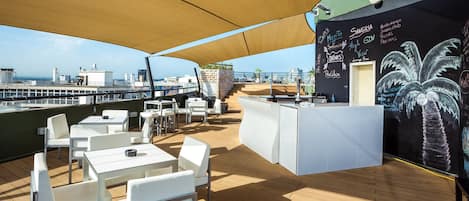 An outdoor deck with white tables and chairs, a bar area with chalkboard menu, and a view of the sea.