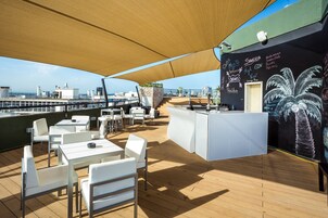 Um deck ao ar livre com mesas e cadeiras brancas, área de bar com cardápio em lousa e vista para o mar.