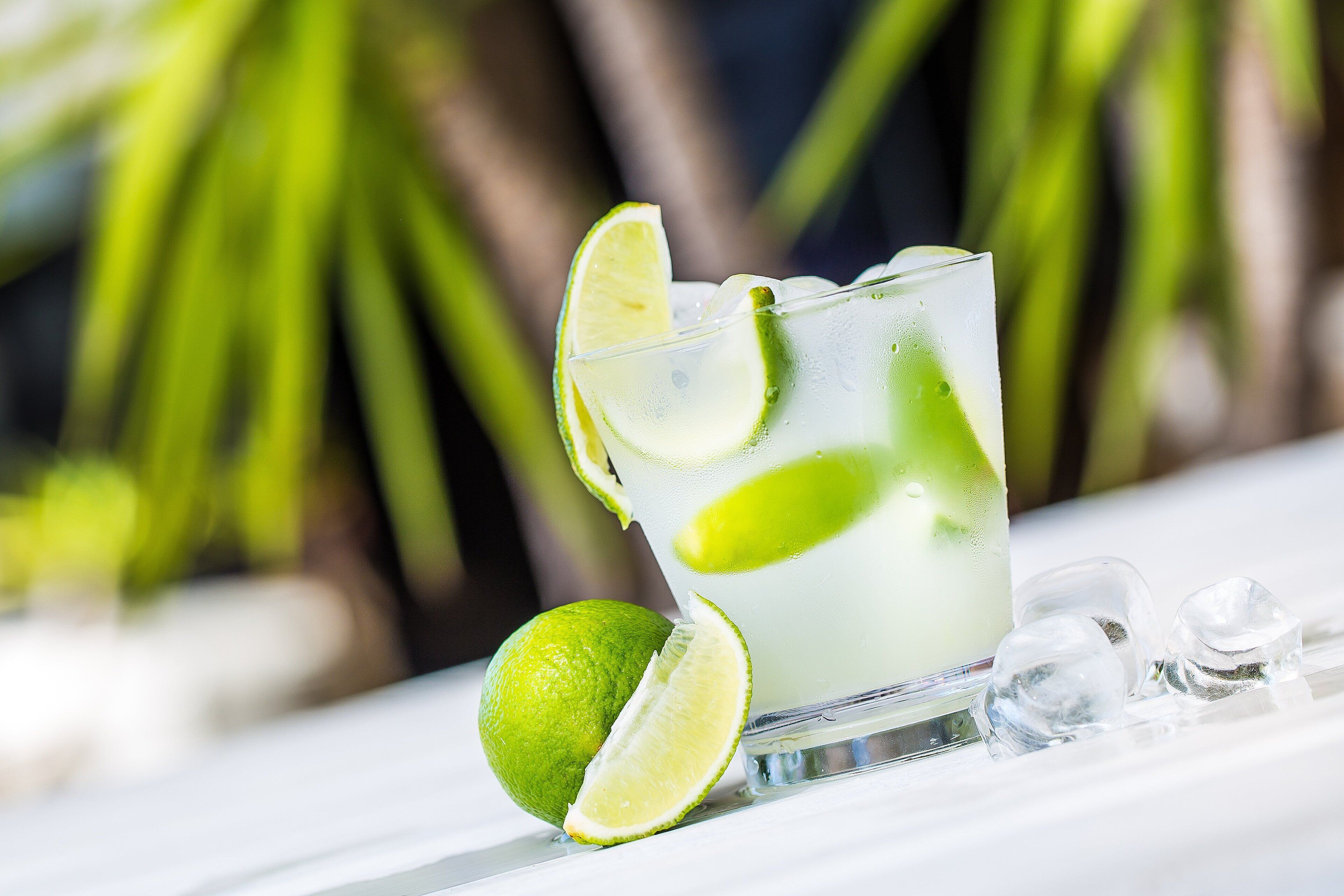 A glass of a clear beverage with lime slices, accompanied by a whole lime and ice cubes on a white surface.