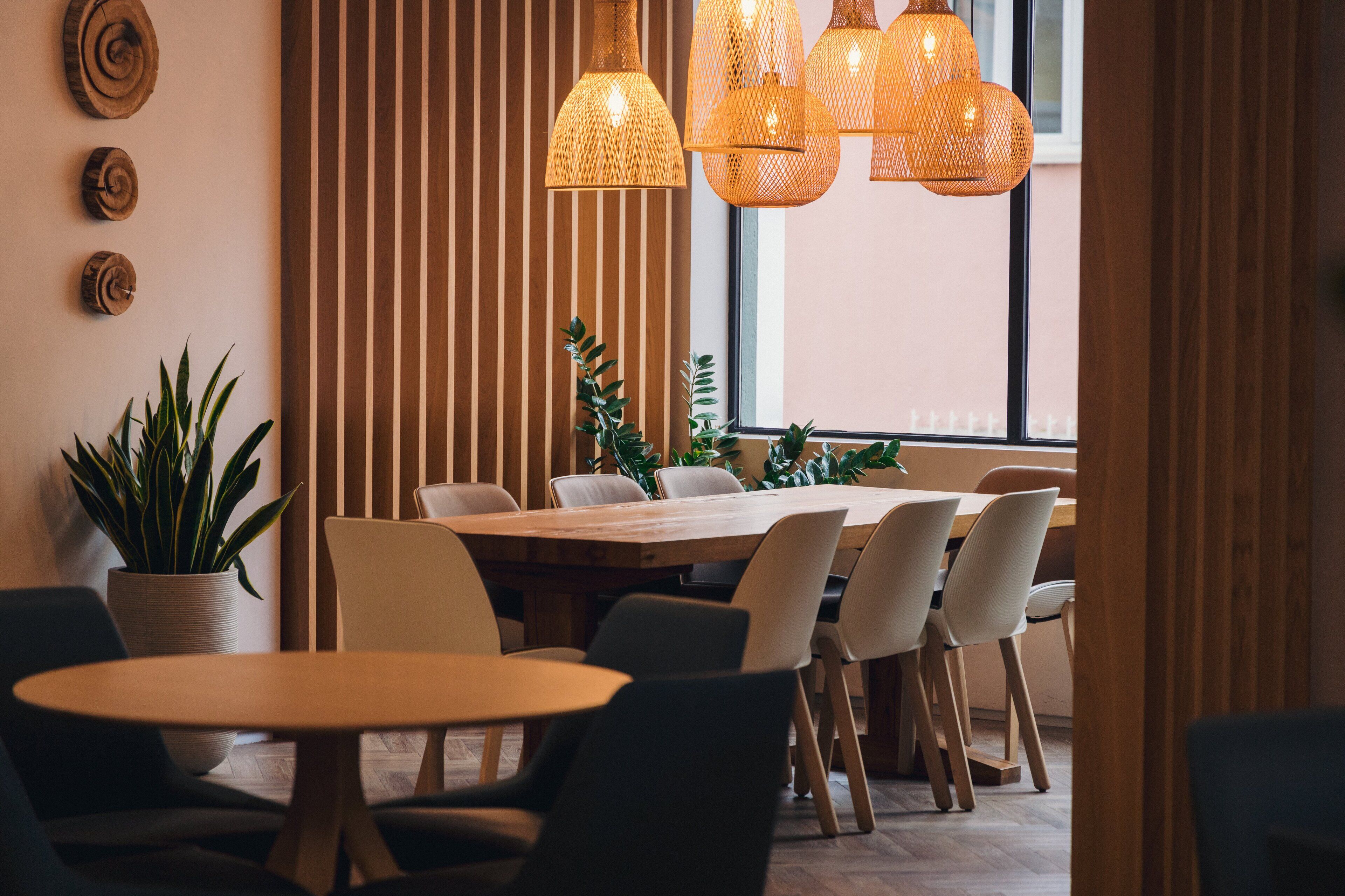 A modern dining area with a wooden table and chairs, potted plants, and woven pendant lights.