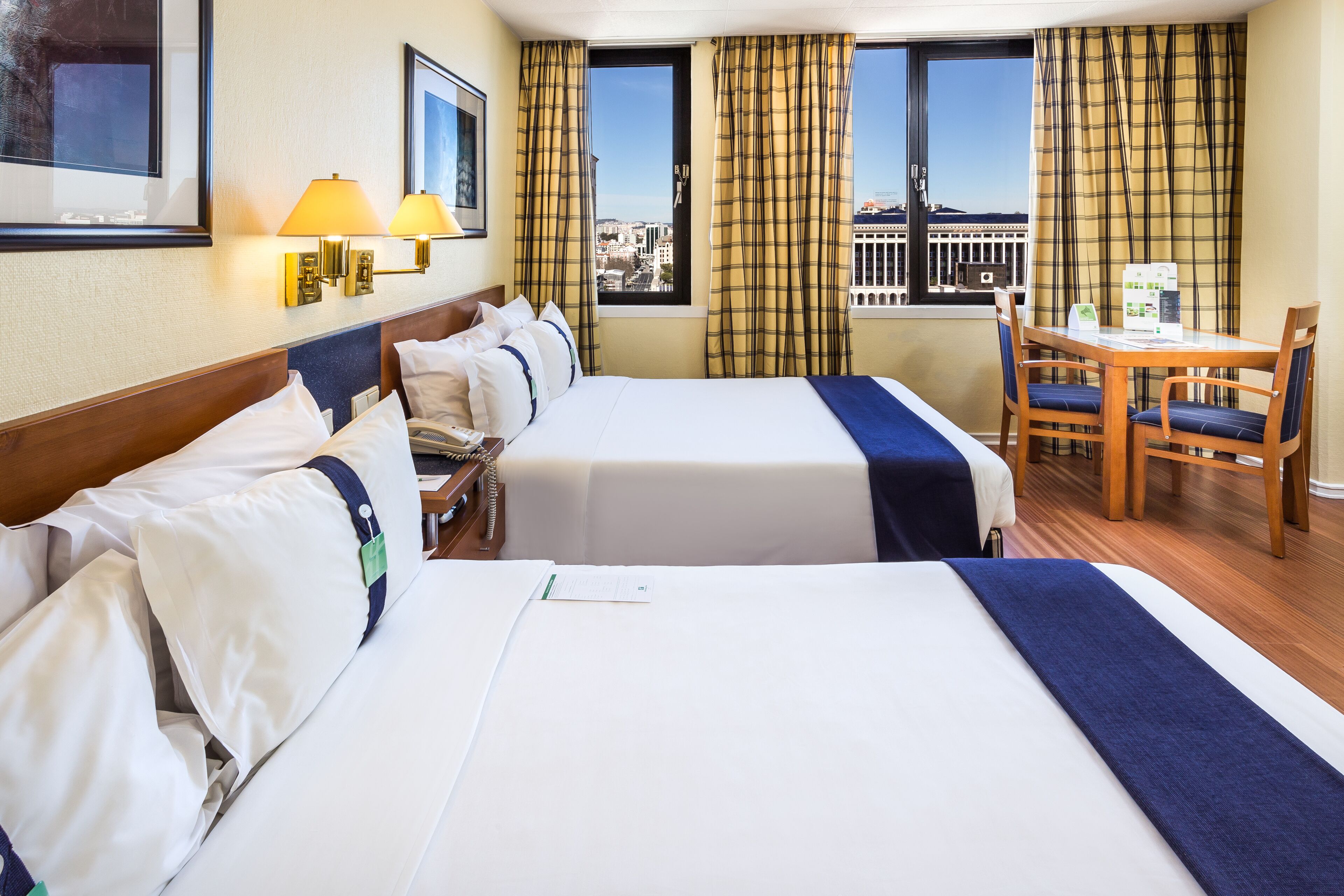 Habitación de hotel con dos camas, un escritorio con sillas, una ventana con cortinas y vista a la ciudad.
