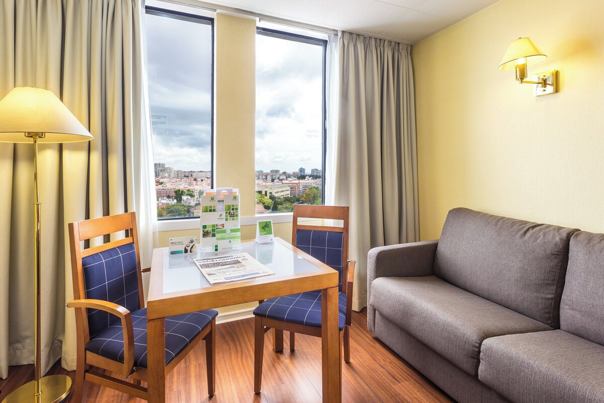 A hotel room with a city view, a dining table with chairs, a sofa, and a lamp.