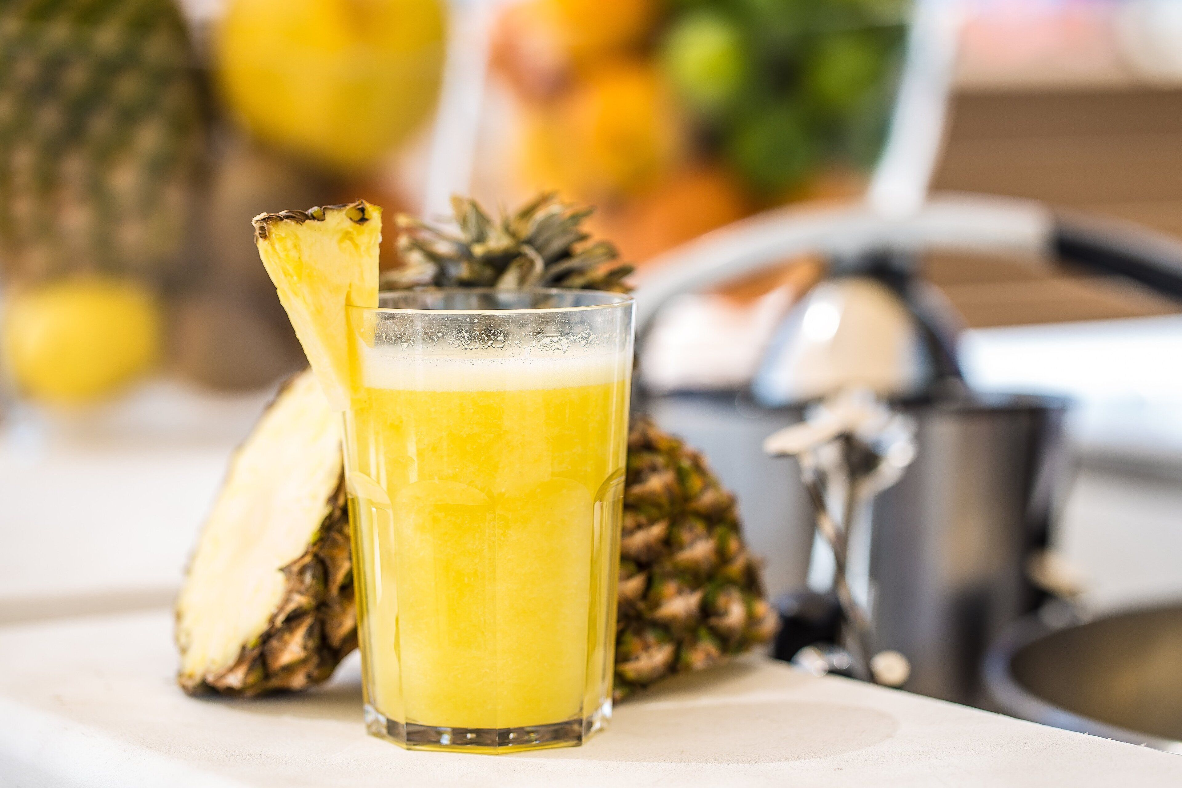 A glass of pineapple juice with a pineapple slice garnish, a whole pineapple, and a metal container in a kitchen setting.