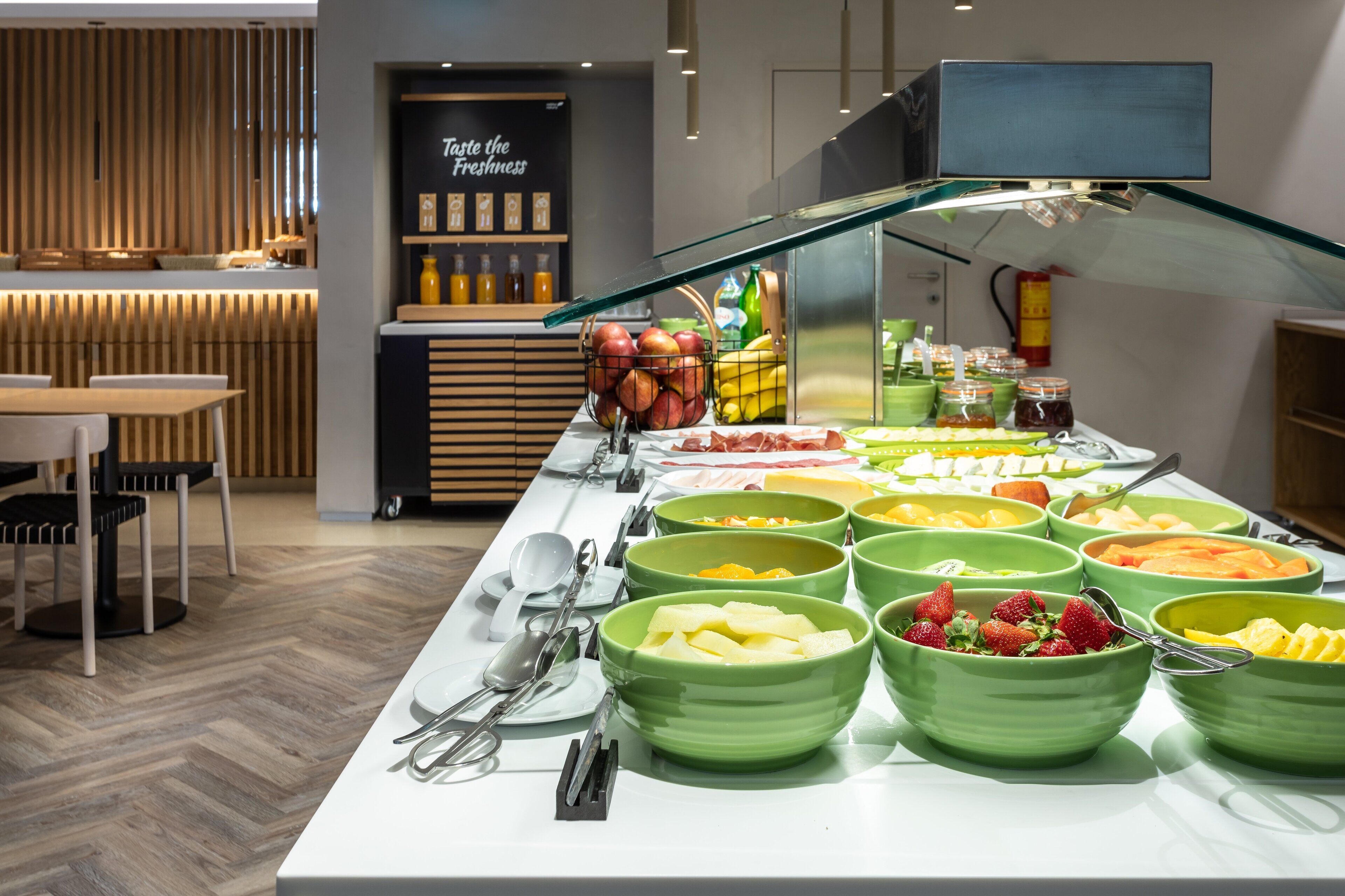 A buffet with various fruits and vegetables in green bowls, a counter with beverages, and a sign that says "Taste the Freshness".