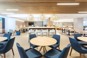 A modern dining area with round tables and blue chairs, a central bar area, and pendant lighting.