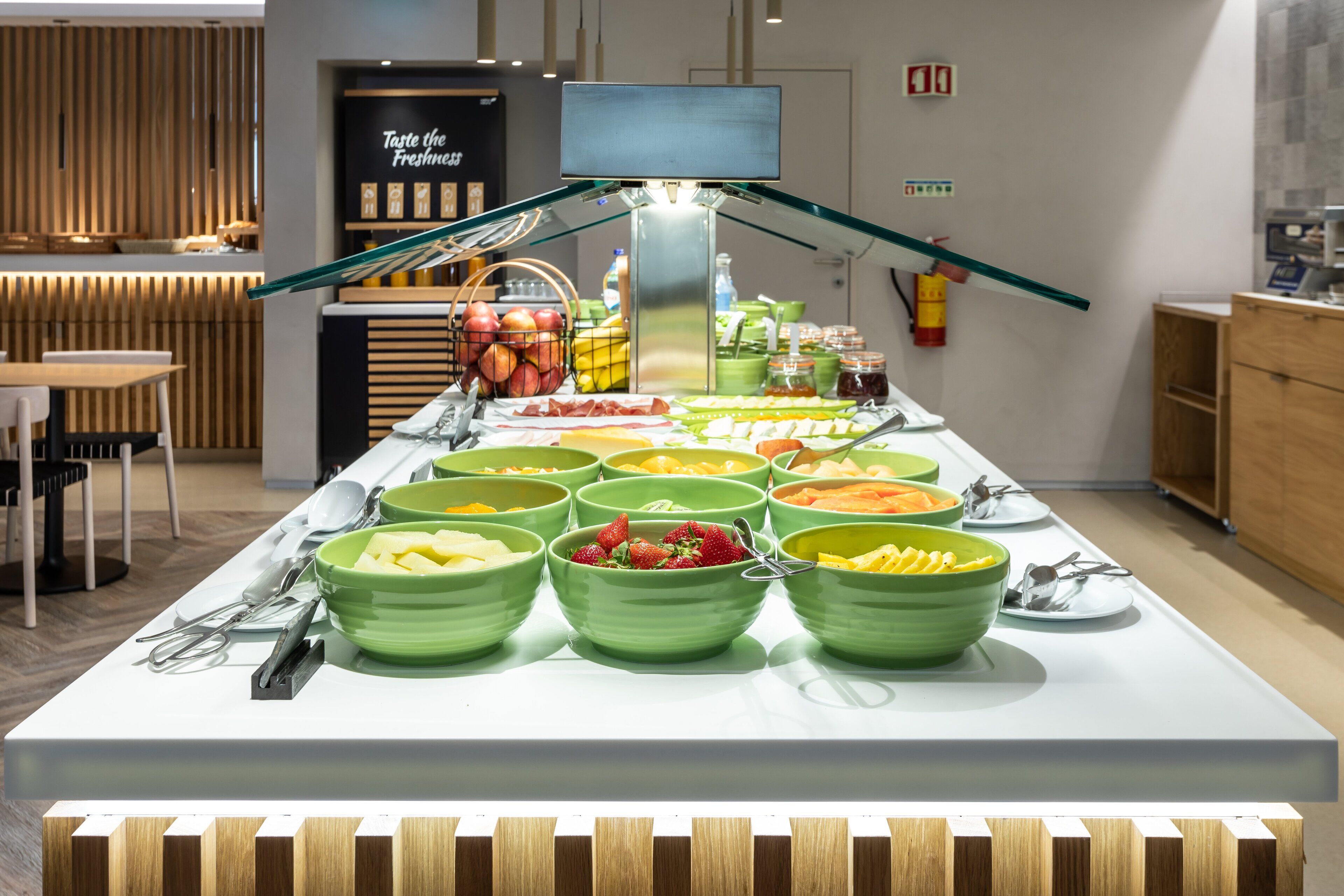 A buffet setup with various food items in green bowls, a television screen, and a wooden wall with a sign.