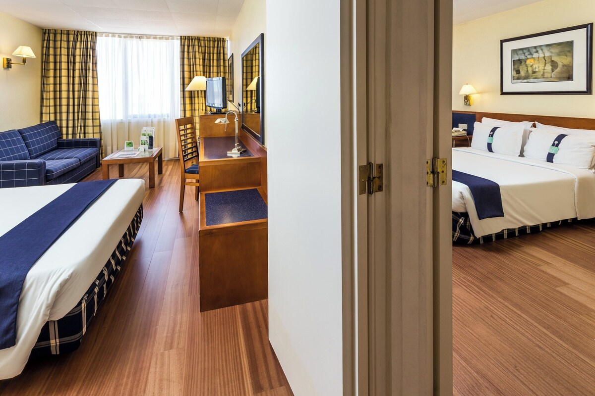 A hotel room with a bed, a sofa, a desk, and a chair. There is a window with curtains, a painting on the wall, and a door.