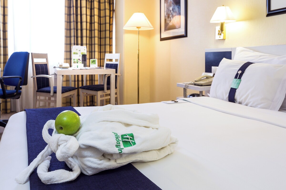 A hotel room with a bed, a desk, chairs, a lamp, and a telephone.