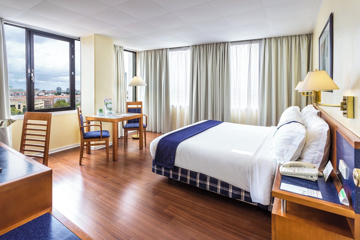 A hotel room with a bed, desk, chairs, and a view of the city.
